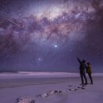 couple stargazing in a beautiful open spot with a blue and purple milkway above them