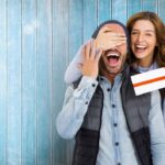 woman giving surprise wrapped gift to man as she covers his eyes with her hand - anniversary gift ideas for him