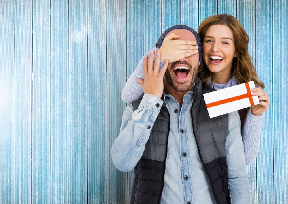 woman giving surprise wrapped gift to man as she covers his eyes with her hand - anniversary gift ideas for him