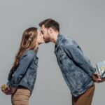 traditional anniversary gifts - image of couple in front of grey background, they are leaning in to kiss on the lips, holding gifts behind their back. both wear denim jackets